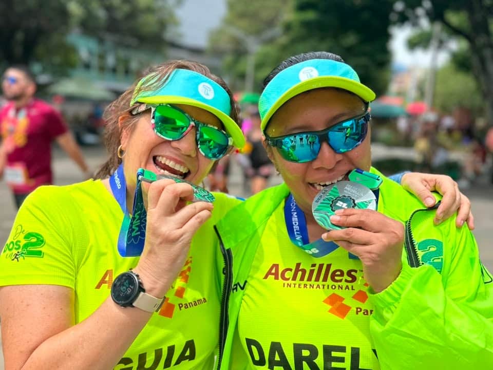 Dos integrantes de la fundación muerden sus medallas al terminar la carrera, una con la camiseta de guía